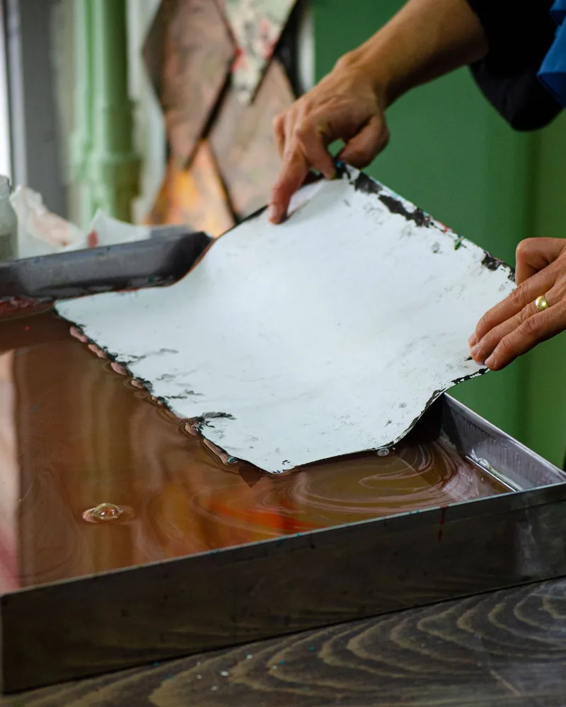 Manos levantando una hoja húmeda de papel sobre una cubeta en una mesa de taller